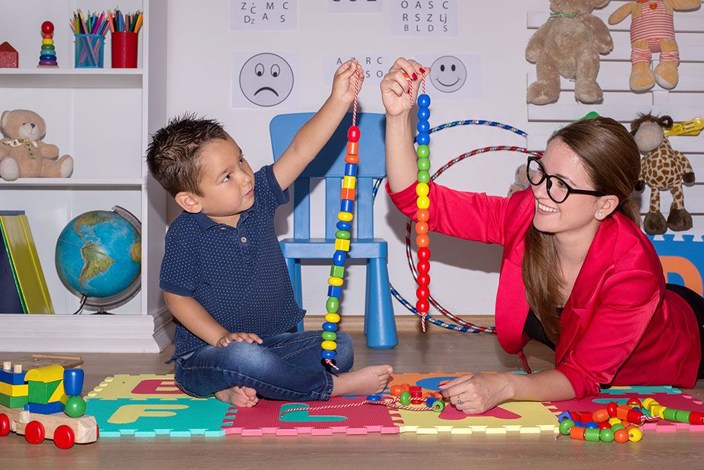 young boy sitting on floor with therapist, each holding a string of beads in the air