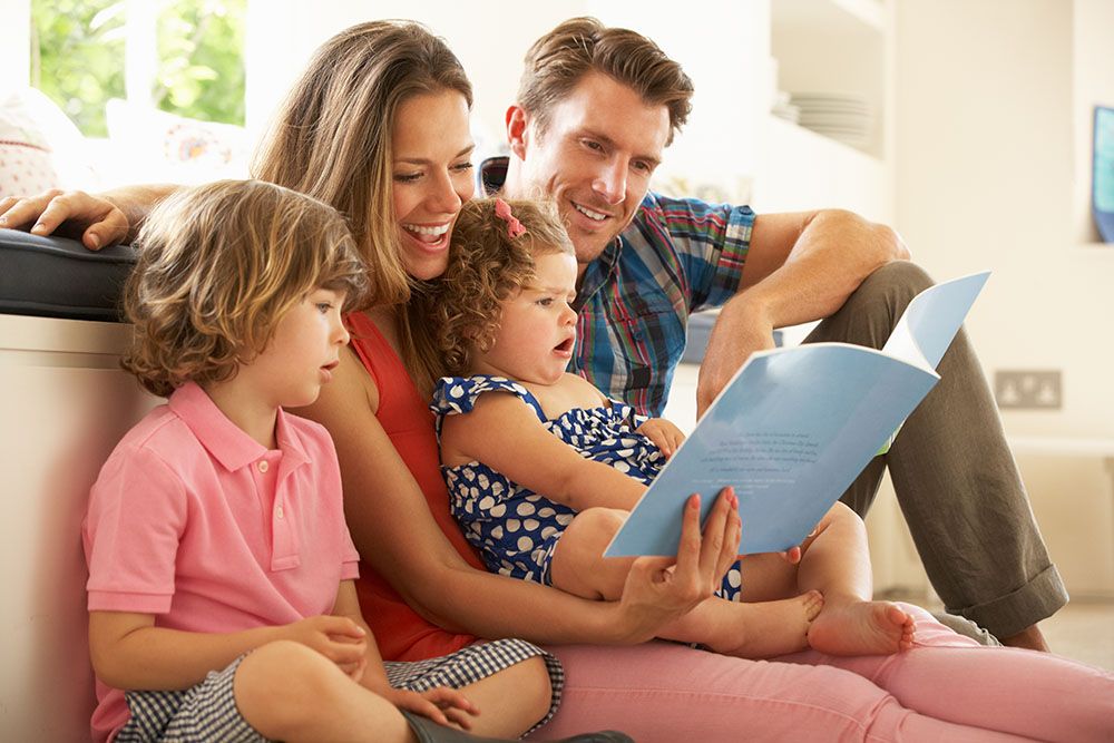 Family sitting on floor with parents reading book to young children.