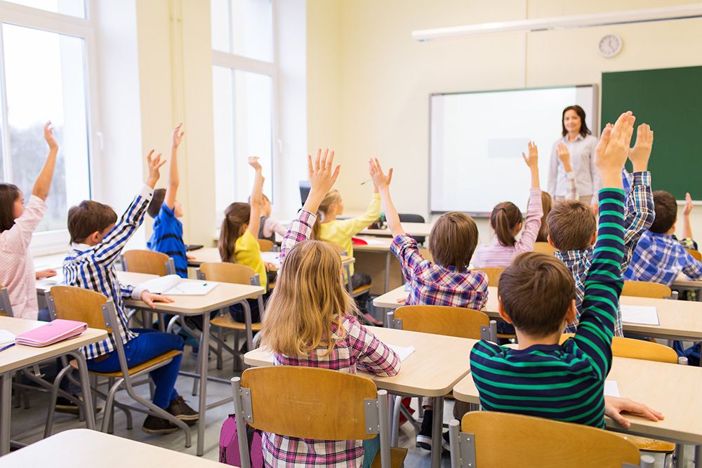 elementary school classroom with teacher up front and all kids raising their hands.